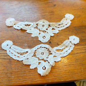 Puede incluir: Dos delicados apliques de encaje blanquecino con diseños florales y circulares. Cada aplique tiene una forma curva con un motivo central y bordes decorativos. El encaje está colocado sobre una superficie de madera.