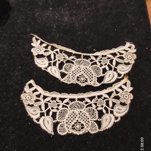 Puede incluir: Dos delicados cuellos de encaje blanco roto con diseños florales y de hojas. El encaje presenta un borde festoneado y se muestra sobre un fondo oscuro. Los cuellos son probablemente vintage o hechos a mano, mostrando una artesanía detallada.