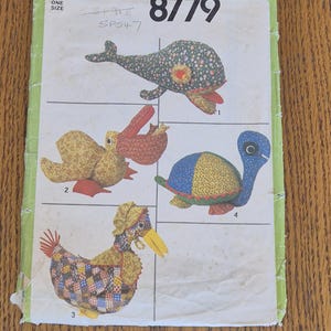 May include: A Simplicity 8779 sewing pattern envelope featuring illustrations of four stuffed animals: a floral whale, a yellow pelican with a red beak, a blue and yellow turtle, and a patchwork chicken wearing a bonnet, all on a light green background.