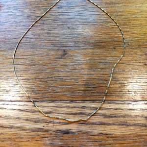 May include: A delicate gold necklace with a fine chain, resting on a wooden surface. The necklace has a simple design with a small clasp. The overall aesthetic is minimalist and elegant, suitable for everyday wear or special occasions.