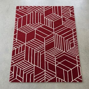 May include: A rectangular rug with a geometric pattern. The rug is primarily a deep red color, with a repeating design of white lines forming three-dimensional cube shapes. The rug is placed on a light gray surface.