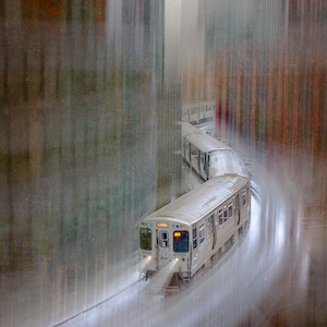 May include: A silver train travels through a city during a rainstorm. The train's headlights illuminate the wet tracks as it curves through the urban landscape. The image has a blurred effect, suggesting movement and the intensity of the rain.