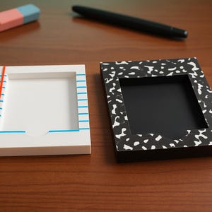 May include: Two rectangular memo holders. One is white with blue horizontal lines and a red vertical line, resembling notebook paper. The other is black with a white speckled pattern, resembling a composition notebook cover. Both have a center cutout.