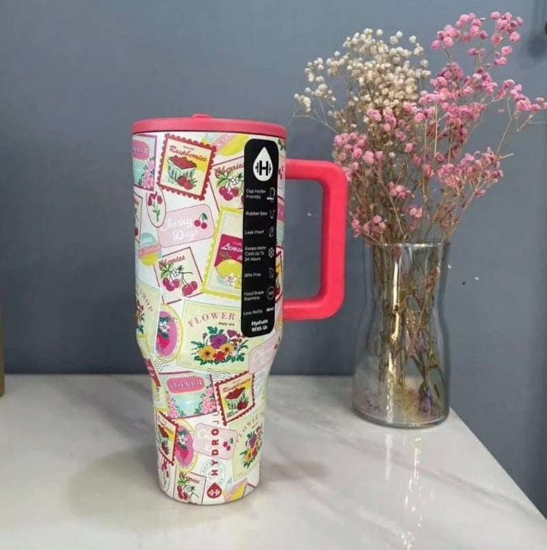 May include: A large, insulated tumbler with a pink handle and lid. The tumbler is covered in a collage of vintage-style fruit and flower labels. A clear vase with pink and white flowers sits to the right.
