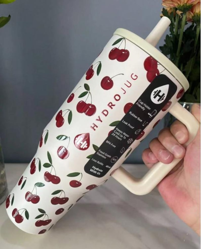 May include: A white Hydrojug with a cherry pattern, a cream-colored handle, and a straw. The jug has the text "HYDROJUG" and other text including "Cup Holder Friendly" and "Keeps Water Cold Up To 24 Hours".