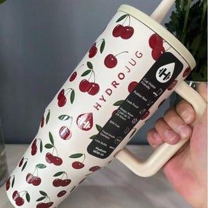 May include: A white Hydrojug with a cherry pattern, a cream-coloured handle, and a straw. The jug has the text "HYDROJUG" and other text including "Cup Holder Friendly", "Leak Proof", and "BPA Free".