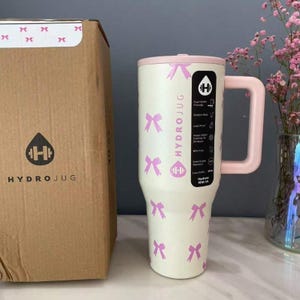 Puede incluir: Una botella de agua HydroJug crema con detalles rosas y un asa. La botella presenta un patr&oacute;n de lazos rosas y el logotipo de HydroJug. A la izquierda hay una caja de cart&oacute;n con el logotipo de HydroJug.