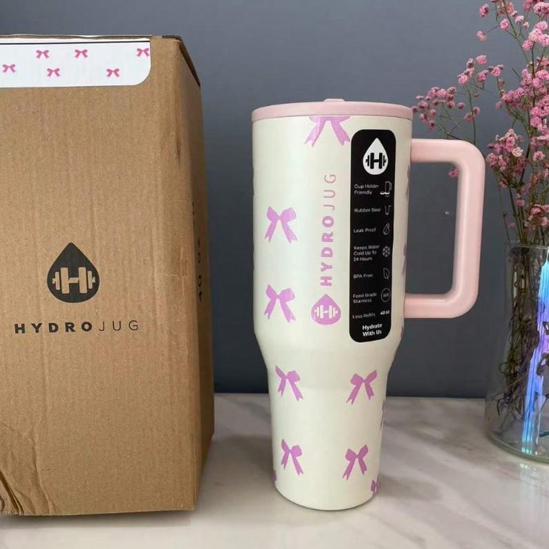 Puede incluir: Una botella de agua HydroJug crema con detalles rosas y un asa. La botella presenta un patr&oacute;n de lazos rosas y el logotipo de HydroJug. A la izquierda hay una caja de cart&oacute;n con el logotipo de HydroJug.