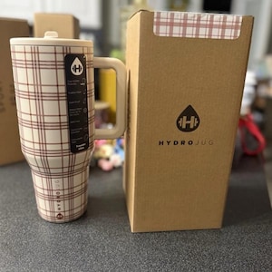 May include: A cream-colored HydroJug with a brown plaid pattern, a handle, and a lid. The HydroJug is next to its cardboard box, which has the HydroJug logo and a plaid pattern on the top. The HydroJug has a capacity of 32 ounces.