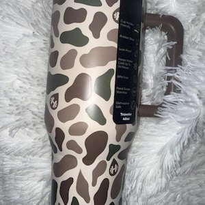 May include: A 40oz insulated travel tumbler with a brown handle and a camouflage pattern in shades of brown, green, and beige. Features include a rubber base, leak-proof design, and BPA-free construction.
