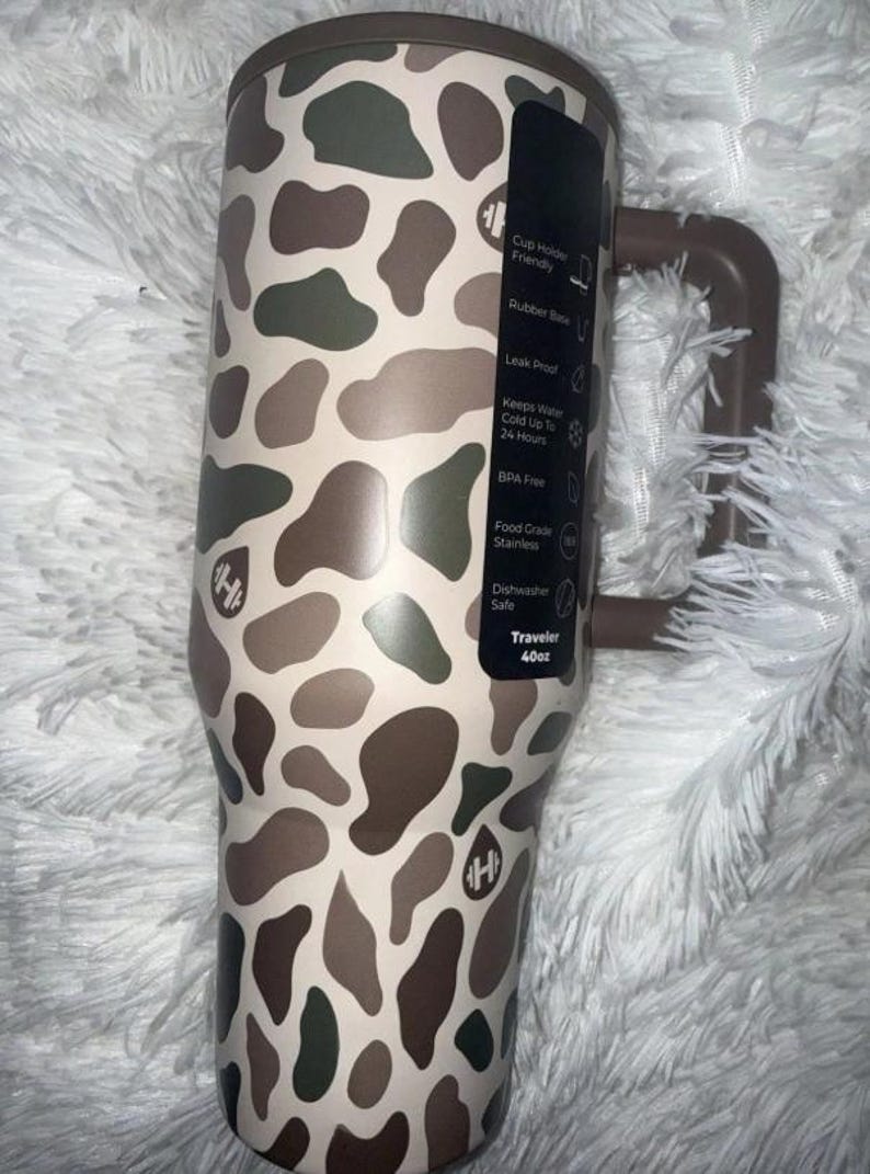 May include: A 40oz insulated travel tumbler with a brown handle and a camouflage pattern in shades of brown, green, and beige. Features include a rubber base, leak-proof design, and BPA-free construction.