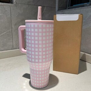 Pu&ograve; includere: Un bicchiere a scacchi rosa e bianco con manico, coperchio e cannuccia. Il bicchiere &egrave; accanto a una scatola di cartone marrone. Il bicchiere &egrave; progettato per contenere una grande quantit&agrave; di liquido.