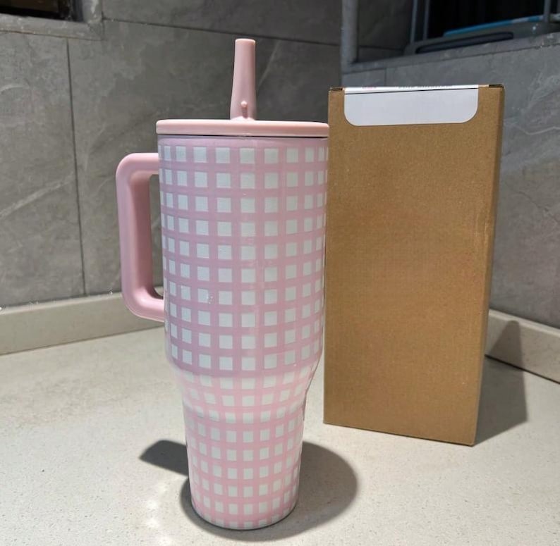 Pu&ograve; includere: Un bicchiere a scacchi rosa e bianco con manico, coperchio e cannuccia. Il bicchiere &egrave; accanto a una scatola di cartone marrone. Il bicchiere &egrave; progettato per contenere una grande quantit&agrave; di liquido.
