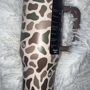 May include: A 40oz insulated travel tumbler with a brown handle and a camouflage pattern in shades of brown, green, and beige. Features include a rubber base, leak-proof design, and BPA-free construction.