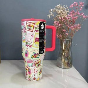 May include: A large, insulated tumbler with a pink handle and lid. The tumbler is covered in a collage of vintage-style fruit and flower labels. A clear glass vase with pink and white flowers sits to the right.