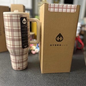 May include: A cream-colored HydroJug with a brown plaid pattern, a handle, and a lid. The HydroJug is next to its cardboard box, which has the HydroJug logo and a plaid pattern on the top. The HydroJug has a capacity of 32 ounces.