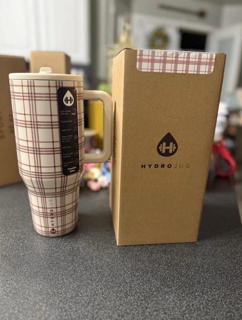 May include: A cream-colored HydroJug with a brown plaid pattern, a handle, and a lid. The HydroJug is next to its cardboard box, which has the HydroJug logo and a plaid pattern on the top. The HydroJug has a capacity of 32 ounces.