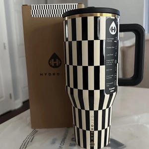 May include: A black and white chequered Hydro Jug travel tumbler with a black handle and lid. The tumbler has a gold rim and the Hydro Jug logo. A brown box with a black and white chequered pattern and the Hydro Jug logo is behind the tumbler.