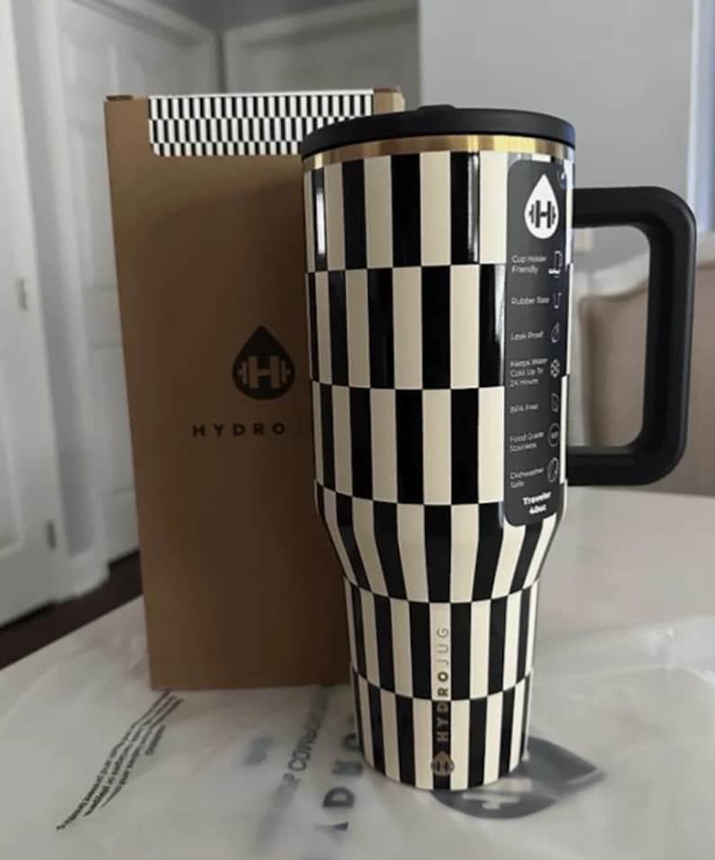 May include: A black and white chequered Hydro Jug travel tumbler with a black handle and lid. The tumbler has a gold rim and the Hydro Jug logo. A brown box with a black and white chequered pattern and the Hydro Jug logo is behind the tumbler.