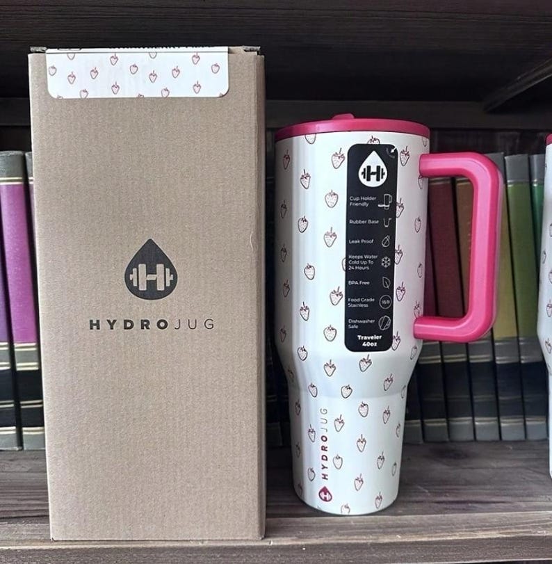 May include: A white HydroJug Traveler 40oz tumbler with a pink handle and lid, adorned with small strawberry illustrations. The tumbler is next to a matching cardboard box with the HydroJug logo.