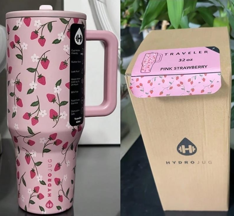 May include: Pink HydroJug water bottle with a handle, featuring a strawberry and floral pattern. The bottle has a lid and a large handle. The packaging label reads "Traveler 32 oz Pink Strawberry". The HydroJug logo is visible.