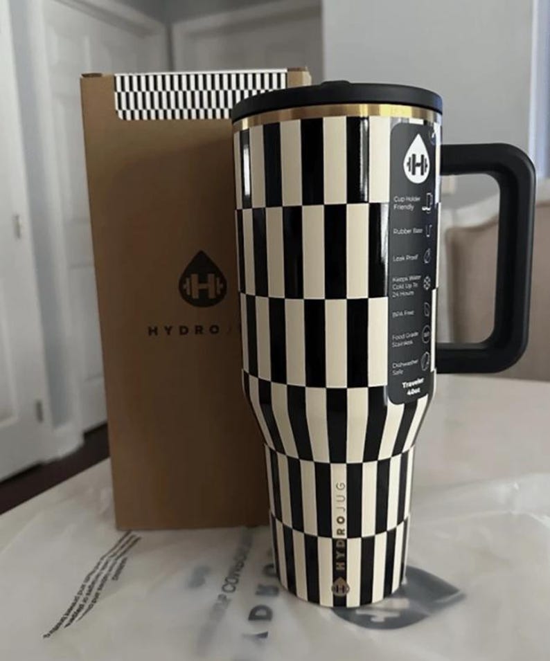 May include: A black and white chequered Hydro Jug travel tumbler with a black handle and lid. The tumbler has a gold rim and the Hydro Jug logo. A brown box with a black and white chequered pattern and the Hydro Jug logo is behind the tumbler.