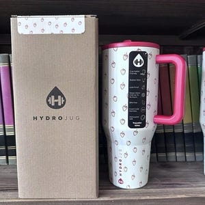 May include: A white HydroJug Traveler 40oz tumbler with a pink handle and lid, adorned with small strawberry illustrations. The tumbler is next to a matching cardboard box with the HydroJug logo.