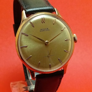 Puede incluir: Un reloj de pulsera Rolex Marconi vintage con esfera y agujas doradas, en una caja dorada. Tiene una correa de cuero negro. La esfera muestra el logotipo de Rolex Marconi.