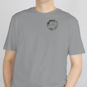 Könnte beinhalten: Ein hellgraues T-Shirt mit Rundhalsausschnitt und kurzen Ärmeln. Ein kreisförmiges Grafikdesign mit den Worten "Quality Driven" und einem grünen Design ist auf der linken Brustseite aufgedruckt.