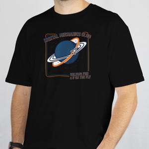 May include: Black t-shirt featuring a graphic of a blue planet with a rocket and the text "ORBITAL MECHANICS CLUB" and "SOLVING FOR AV ON THE FLY" in white and orange. The design is enclosed in a rectangular orange border.
