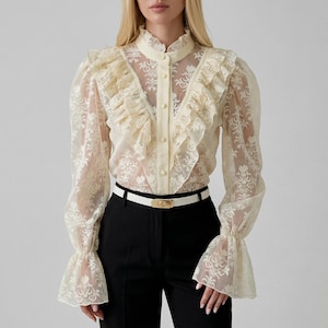 Puede incluir: Blusa de encaje color crema con cuello alto, volantes en la parte delantera y mangas largas. La blusa tiene una botonadura frontal y se combina con pantalones negros y un cinturón blanco con hebilla dorada. El patrón floral de encaje añade un toque vintage.
