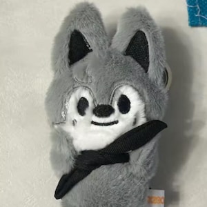 May include: A plush gray wolf keychain with black accents on the ears and a black bow tie. The wolf has a white face with black eyes and a small black nose. The keychain is made of soft, fuzzy material.