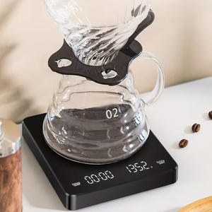 May include: A clear glass coffee pour-over set with a black plastic holder and a black digital scale. The scale displays time and weight. Coffee beans are scattered around the set.