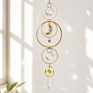 May include: A hanging suncatcher featuring gold-toned metal rings, two clear faceted crystal spheres, and a clear crystal point. It includes a crescent moon charm with blue floral details, a small blue bead, and a gold disc with baby footprint engravings.