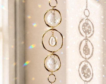 May include: A hanging suncatcher featuring a gold-toned chain with three interconnected gold rings. Two rings contain clear, faceted spherical crystals, while the center ring holds a clear, teardrop-shaped crystal. A small gold wing charm hangs at the bottom.