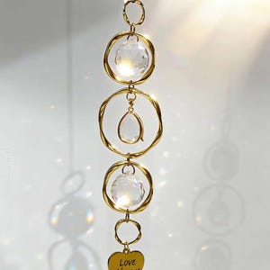 Könnte beinhalten: Ein dekorativer goldfarbener Sonnenfänger mit klaren Kristallkugeln und einem tropfenförmigen Kristall. Ein goldener Herzanhänger am unteren Ende trägt die Aufschrift "Love Always". Der Sonnenfänger hängt an einer filigranen Kette.