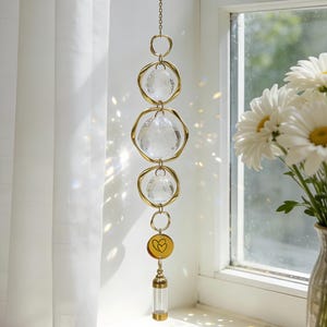 May include: A decorative suncatcher with clear crystal spheres and gold-coloured metal accents. The suncatcher hangs from a chain and features a small, round charm with a heart design and a small glass vial. The suncatcher is hanging in front of a window.