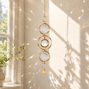 May include: A gold-toned hanging suncatcher featuring a vertical chain with three circular gold rings, two faceted clear crystal spheres, one central circular mirror, and a small gold disc at the bottom engraved with the letter A.