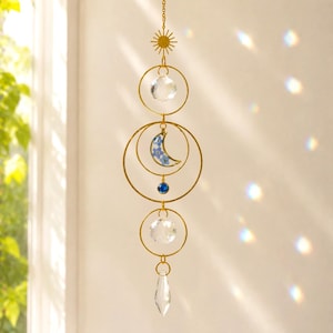 Può includere: Un acchiappasole sospeso color oro con un ciondolo a forma di sole in alto, due sfere di cristallo sfaccettato trasparente, un ciondolo a mezzaluna con piccoli fiori blu, una perlina blu e un cristallo trasparente a goccia in fondo.