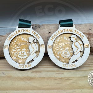 Puede incluir: Dos medallas circulares de madera con el texto MULLERTHAL TRAIL y 112 KM. Cada medalla muestra una escena grabada con láser de un corredor, un puente de piedra y una cascada. Las medallas tienen una cinta verde oscuro y la letra M en los laterales.