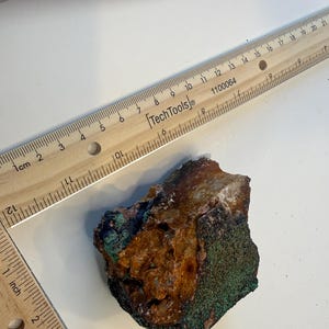 May include: A rough-hewn rock sample with a mix of brown, green, and black colors. The rock is next to a wooden ruler marked in inches and centimeters. The ruler is labeled "Tech Tools".