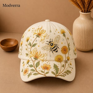 Puede incluir: Una gorra de béisbol de color crema con un diseño bordado de una abeja rodeada de flores de diente de león amarillas y hojas verdes. La gorra está adornada con pequeños diamantes de imitación transparentes dispersos entre los motivos.