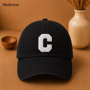 Op de afbeelding: Een zwarte baseballpet met een grote, witte, geborduurde letter C op het voorpaneel. De pet heeft een gestructureerde kroon, een gebogen klep en zichtbare stiksels. De tekst Modverra staat linksboven afgedrukt.