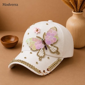 Puede incluir: Una gorra de béisbol blanca con una gran aplicación de mariposa hecha de lentejuelas iridiscentes rosas y amarillas, pequeñas perlas blancas y un detalle de flor rosa. La visera está decorada con pedrería dorada y tres anillas de metal dorado.