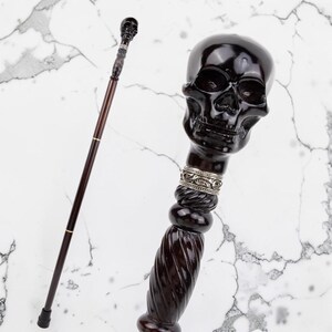 Pode incluir: Uma bengala castanha escura com uma pega em forma de caveira. A bengala apresenta um design detalhado de caveira, uma faixa prateada e uma pega inferior em espiral. O eixo é castanho escuro com um detalhe dourado. A bengala tem uma ponta de borracha.