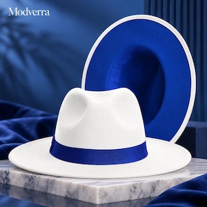 May include: A white fedora hat with a wide brim and a vibrant blue ribbon band. The text Modverra is visible in the upper left corner. A second hat is positioned behind the first, showing a blue underside with a white brim edge.