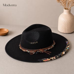 May include: A black wide-brimmed hat featuring a pinched crown with five embroidered tan X stitches. The base of the crown is wrapped in a brown leather cord and a multi-colored patterned fabric trim that extends onto the brim's edge.