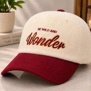 Op de afbeelding: Een crèmekleurige corduroy baseballpet met een donkerrode klep. De voorkant is voorzien van geborduurde tekst in donkerrood met de woorden BE WILD AND in een eenvoudig lettertype en het woord Wonder in een groter cursief schrift met gele rand.