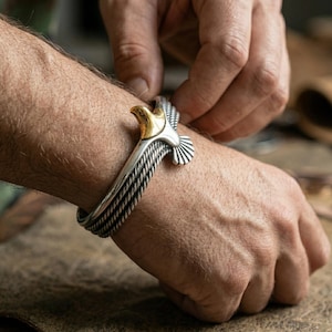 Puede incluir: Brazalete plateado con la cabeza de un águila detallada con detalles dorados. El brazalete tiene un diseño de cuerda trenzada. La cabeza del águila está en la parte superior del brazalete, con las alas extendidas.