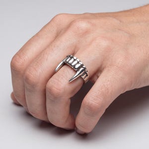 May include: A silver-colored ring designed to resemble a set of vampire fangs. The ring features two prominent, pointed fangs on either side, with a row of smaller teeth in between. The ring is worn on a finger, showcasing its gothic and unique design.